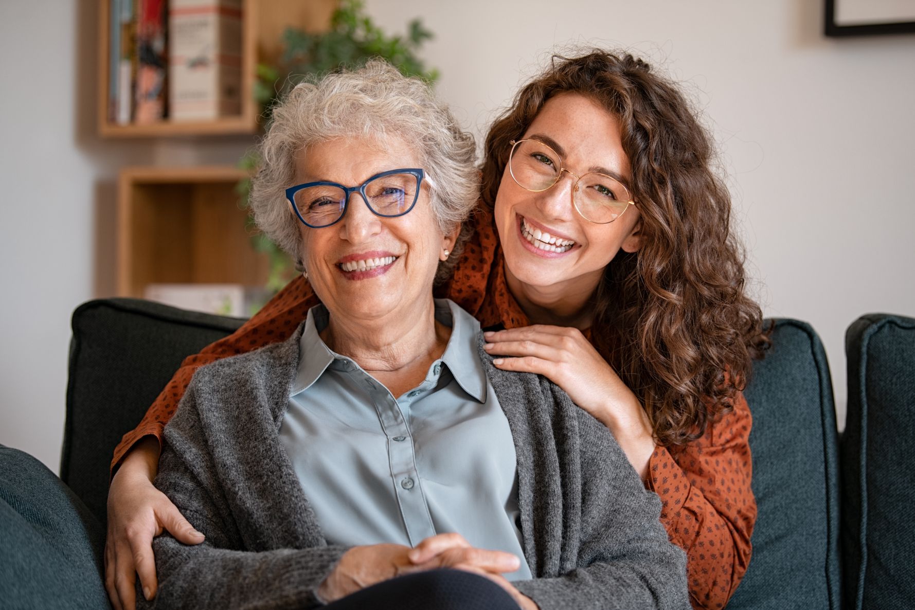 Portrait of old grandma and adult granddaughter hugging with love on sofa while looking at camera. Happy young woman with eyeglasses hugging from behind older grandma with spectacles. Senior woman spending time with her beautiful daughter, generation family concept.